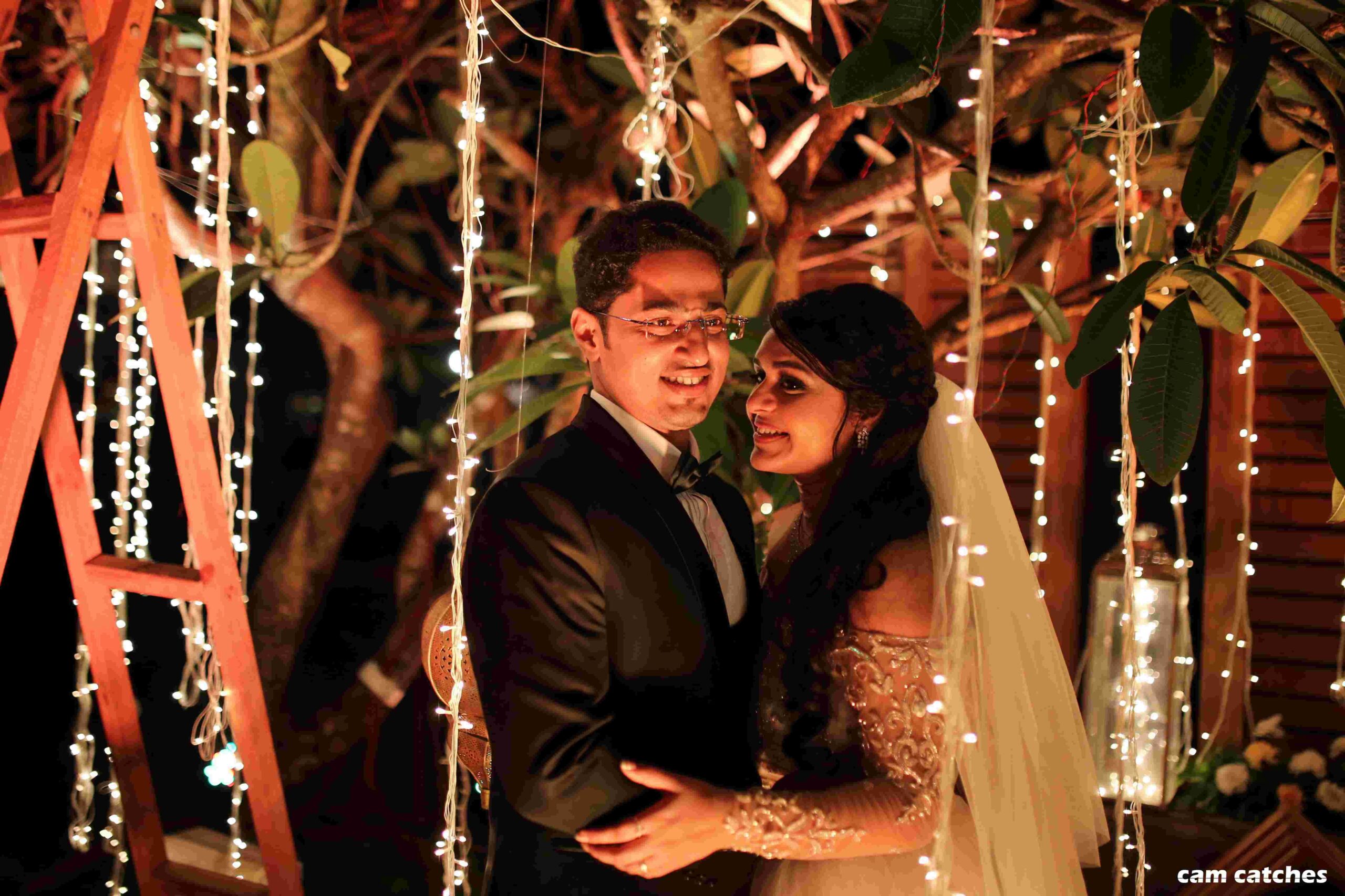 A romantic wedding reception scene with a smiling bride and a happy groom embracing under cascading white lights holding each other.