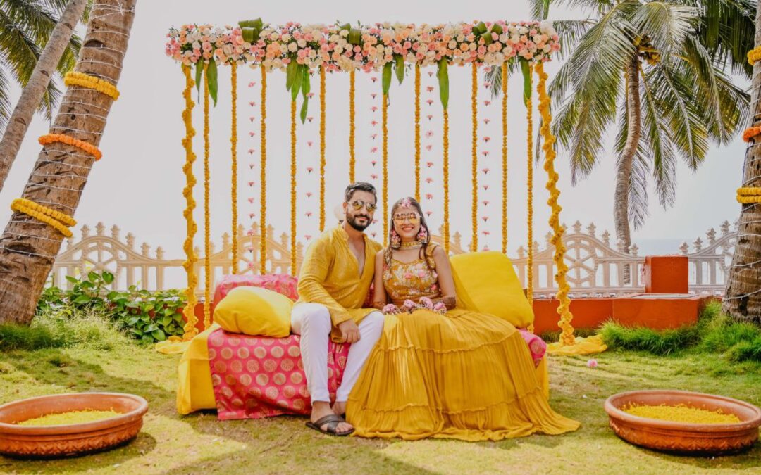 A vibrant outdoor haldi ceremony scene with a bride and groom seated on a cushioned seating arrangement, surrounded by floral decorations, palm trees, and a festive, tropical atmosphere.