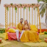 A vibrant outdoor haldi ceremony scene with a bride and groom seated on a cushioned seating arrangement, surrounded by floral decorations, palm trees, and a festive, tropical atmosphere.