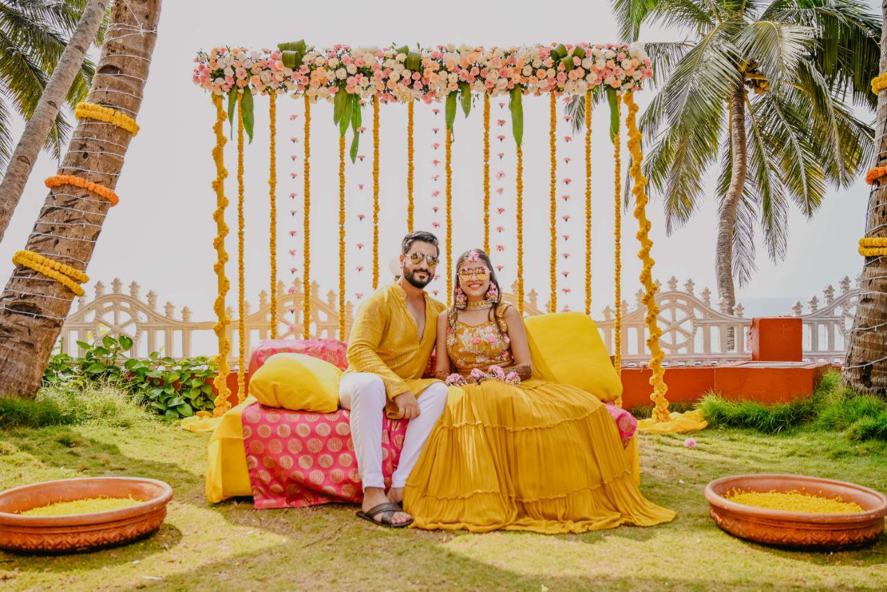 A vibrant outdoor haldi ceremony scene with a bride and groom seated on a cushioned seating arrangement, surrounded by floral decorations, palm trees, and a festive, tropical atmosphere.