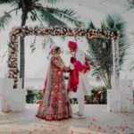 Newlyweds exchange varmala under a floral archway with a stunning ocean view.