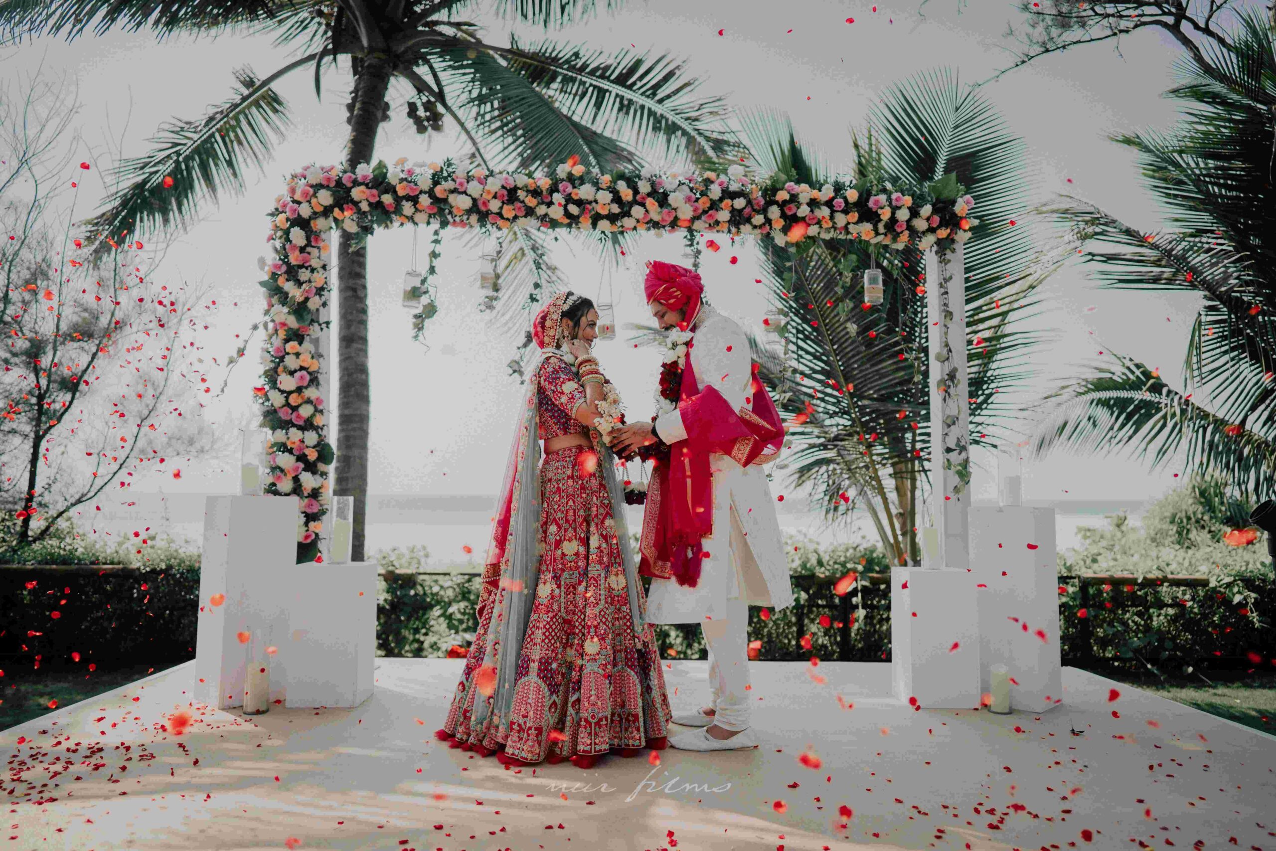 Newlyweds exchange varmala under a floral archway with a stunning ocean view.