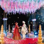 A newly wedded stand beneath a flower canopy holding hands.