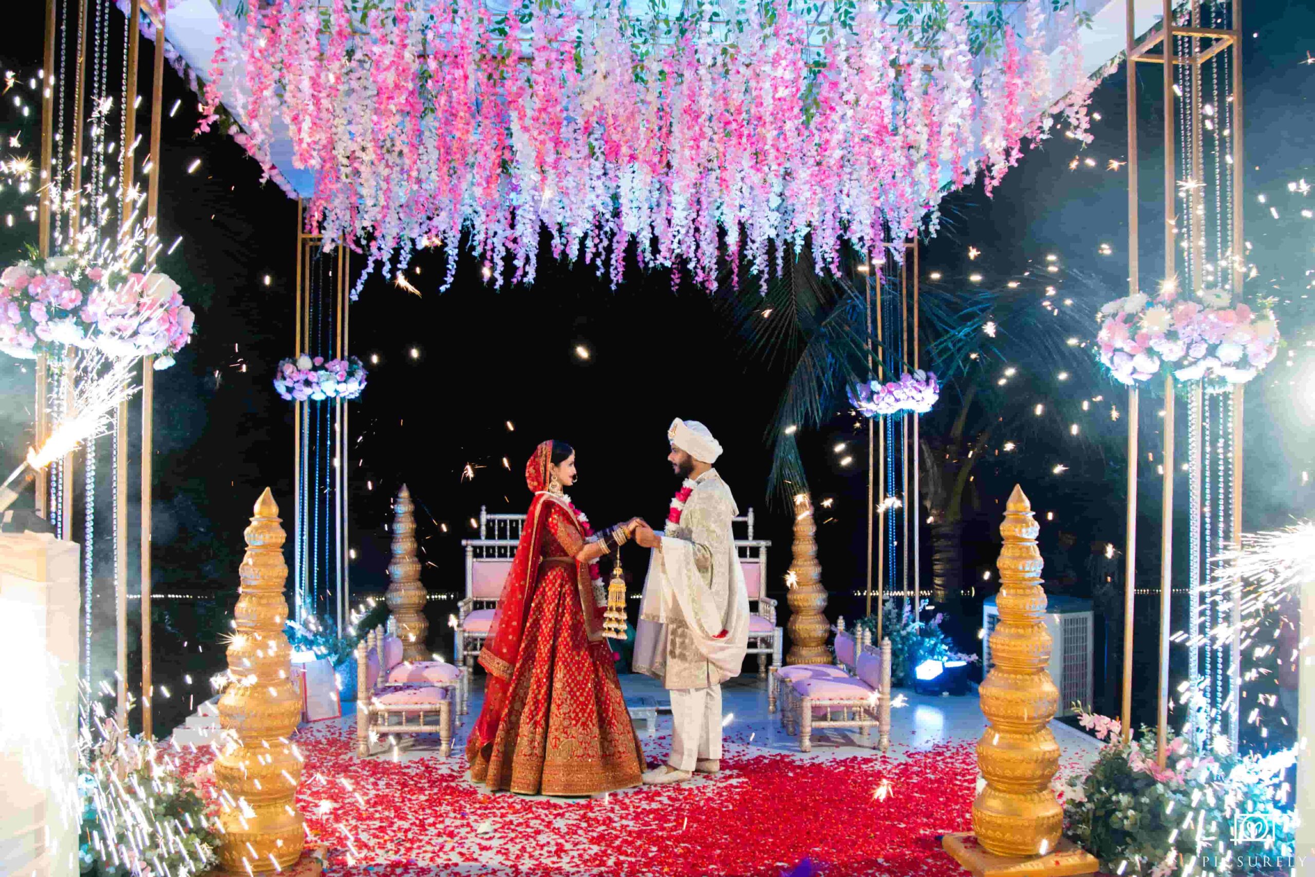 A newly wedded stand beneath a flower canopy holding hands.